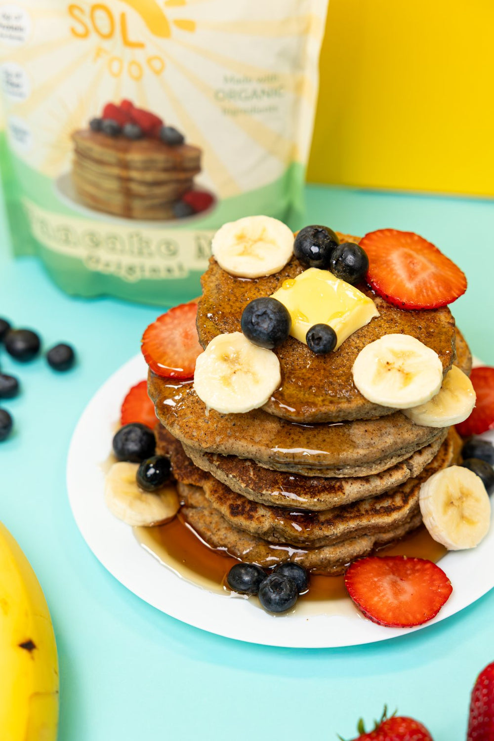 A tall stack of Sol Food pancakes topped with banana, strawberries, blueberries and syrup