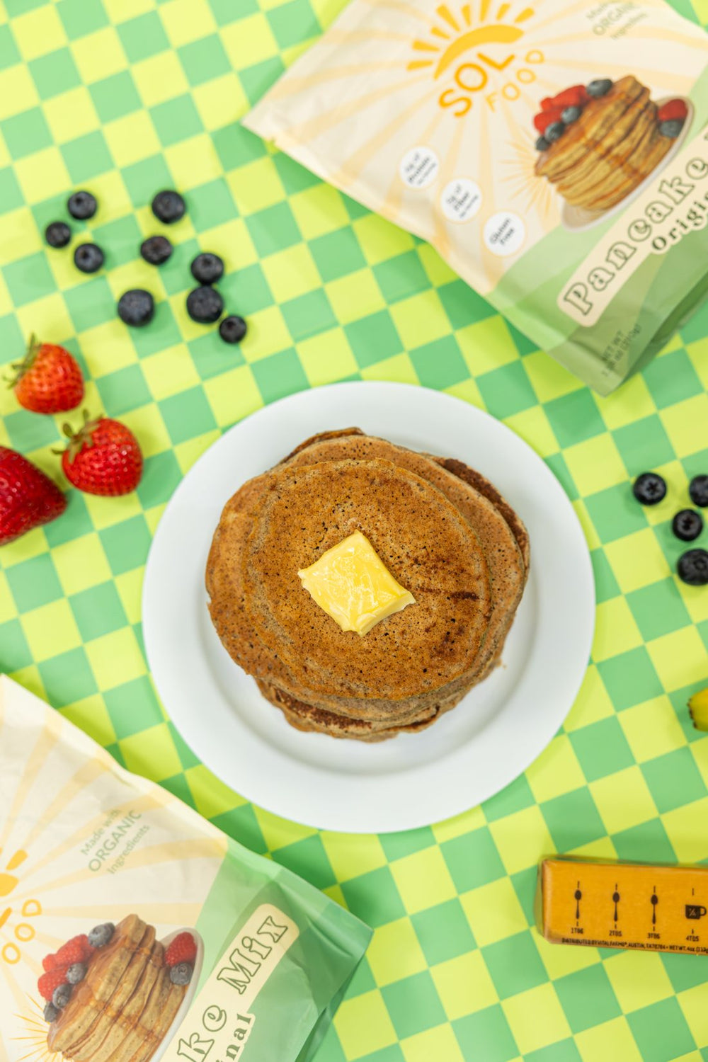 A stack of Sol Food pancakes with butter on a green checkered table, next to two bags of Original Pancake Mix